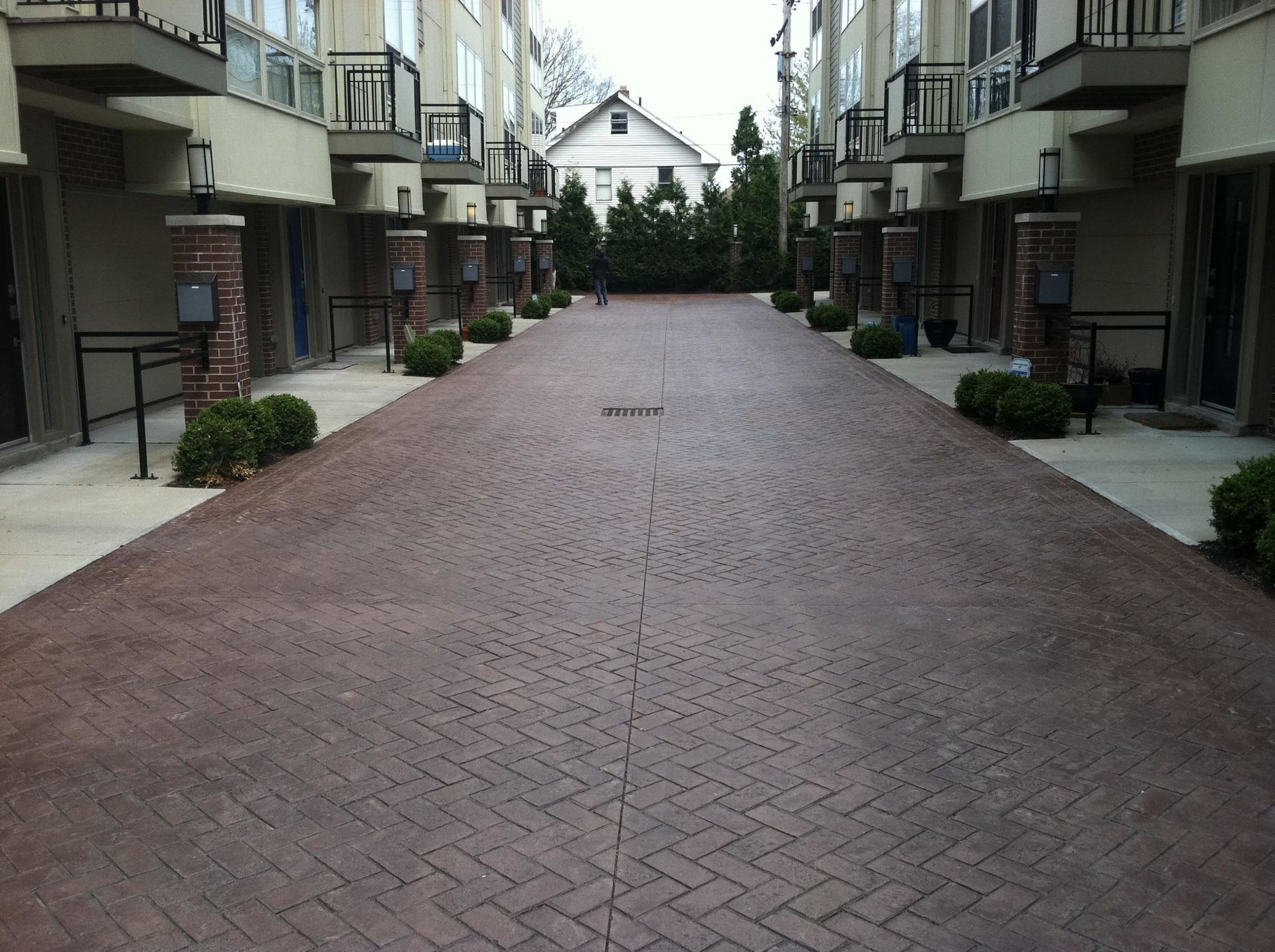 Brick-paved alleyway between townhouses with mailboxes, bushes, and a distant house.
