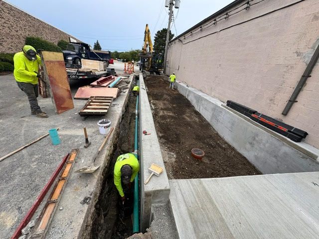 Construction workers in neon vests install a pipe in a trench next to a building.
