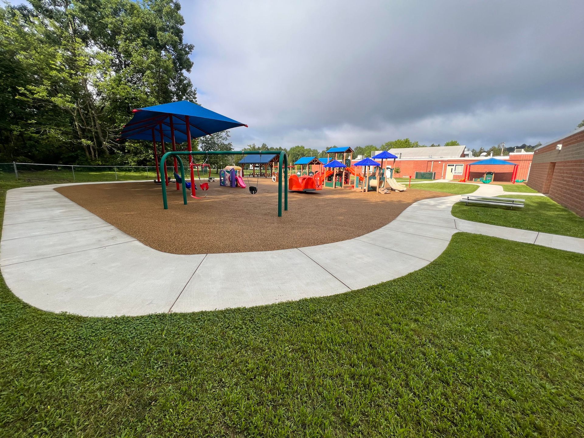Playground with swings and climbing structures on wood chips, surrounded by grass and a sidewalk.