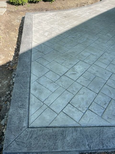 Freshly poured concrete slab with diagonal stamped pattern and dark border beside bare soil.