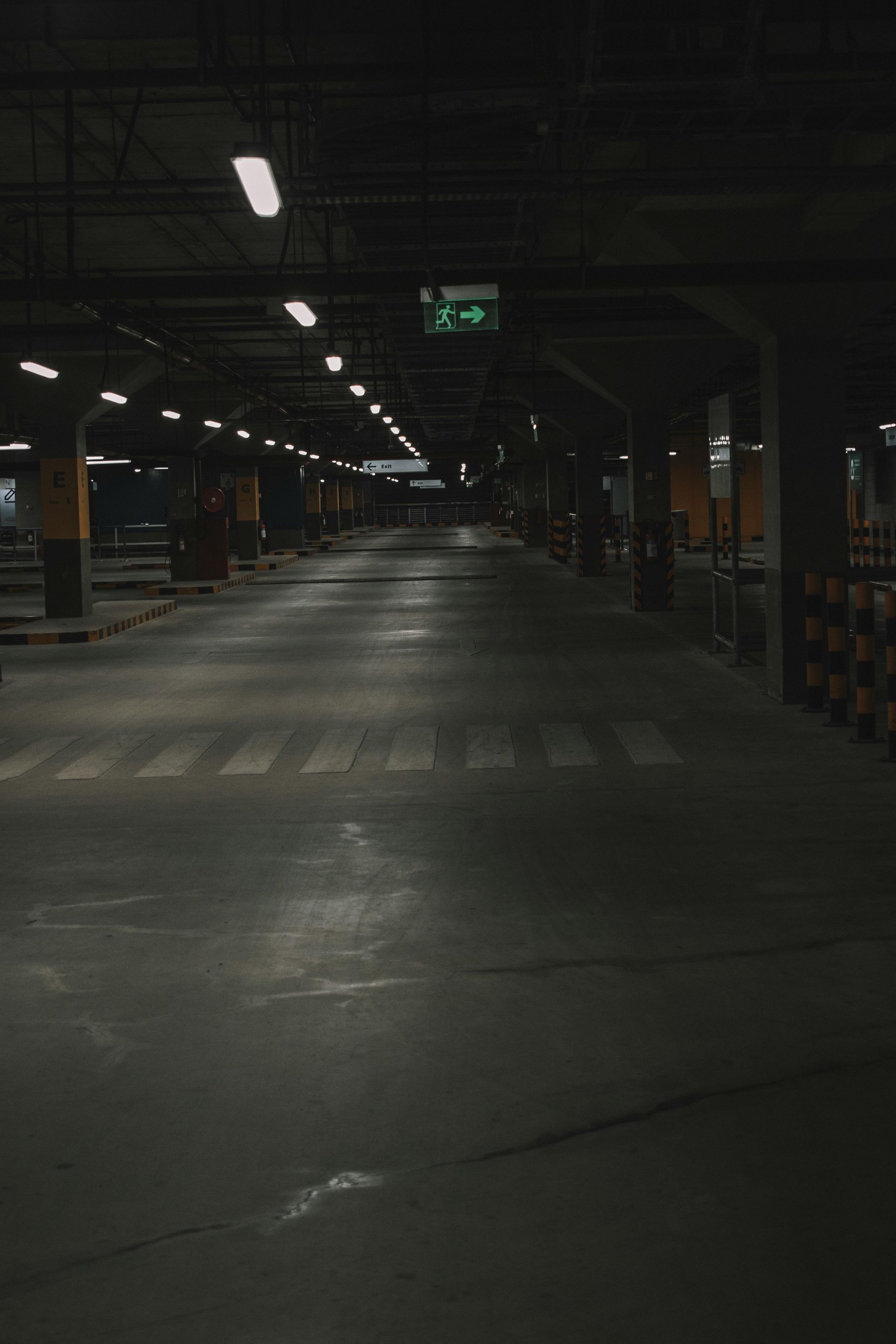 Dark, empty parking garage with overhead lights. Exit sign glowing green at the end of the aisle.