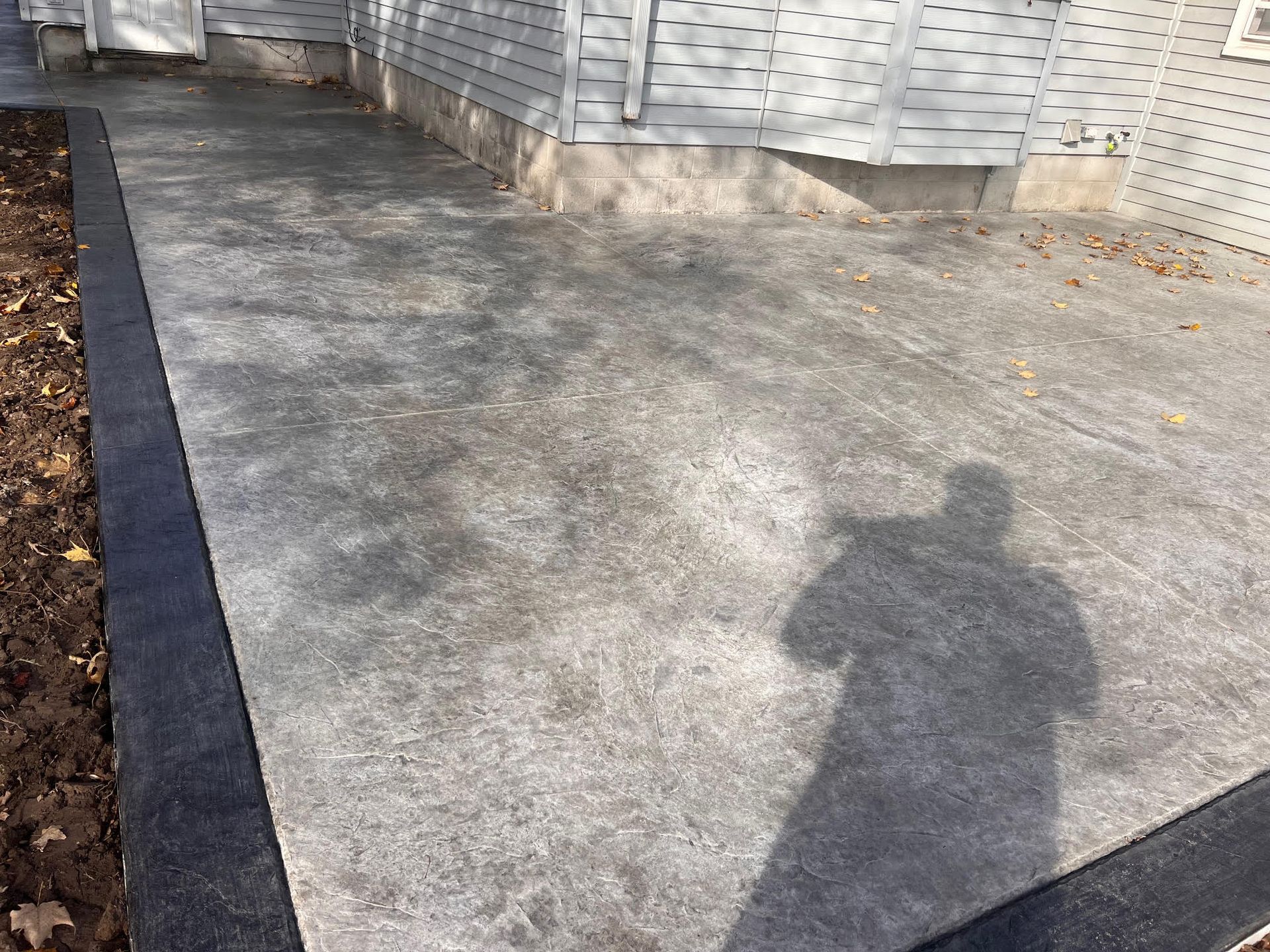 Concrete patio with dark border next to a house with beige siding and a shadow of a person.