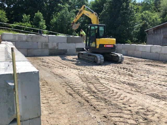 Excavator inside a square of concrete blocks on a sandy surface, under a blue sky.