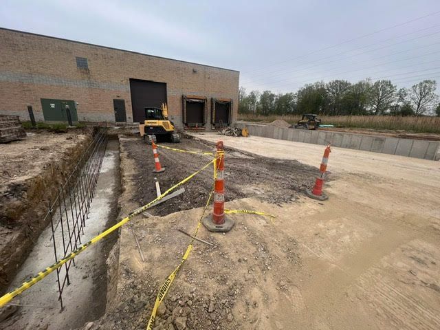 Construction site: Excavated trench with rebar, caution tape, orange cones, and a small vehicle near a loading dock.