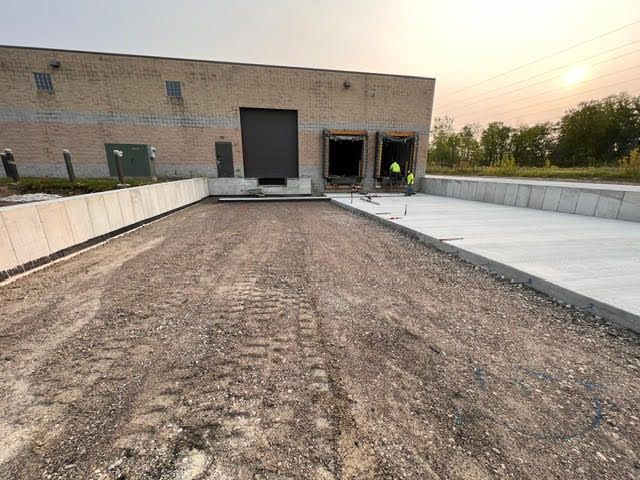Exterior view of a loading dock area; a brick building with two open bays, workers present, gravel driveway.
