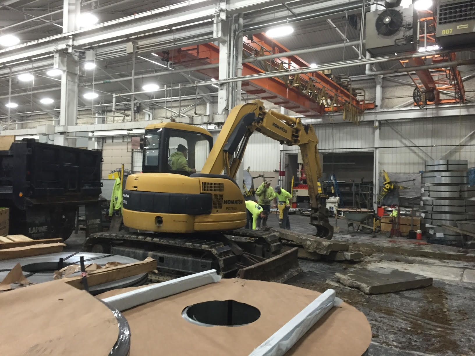 Yellow excavator removing debris in a factory; workers in safety vests nearby; metal coils and overhead crane visible.