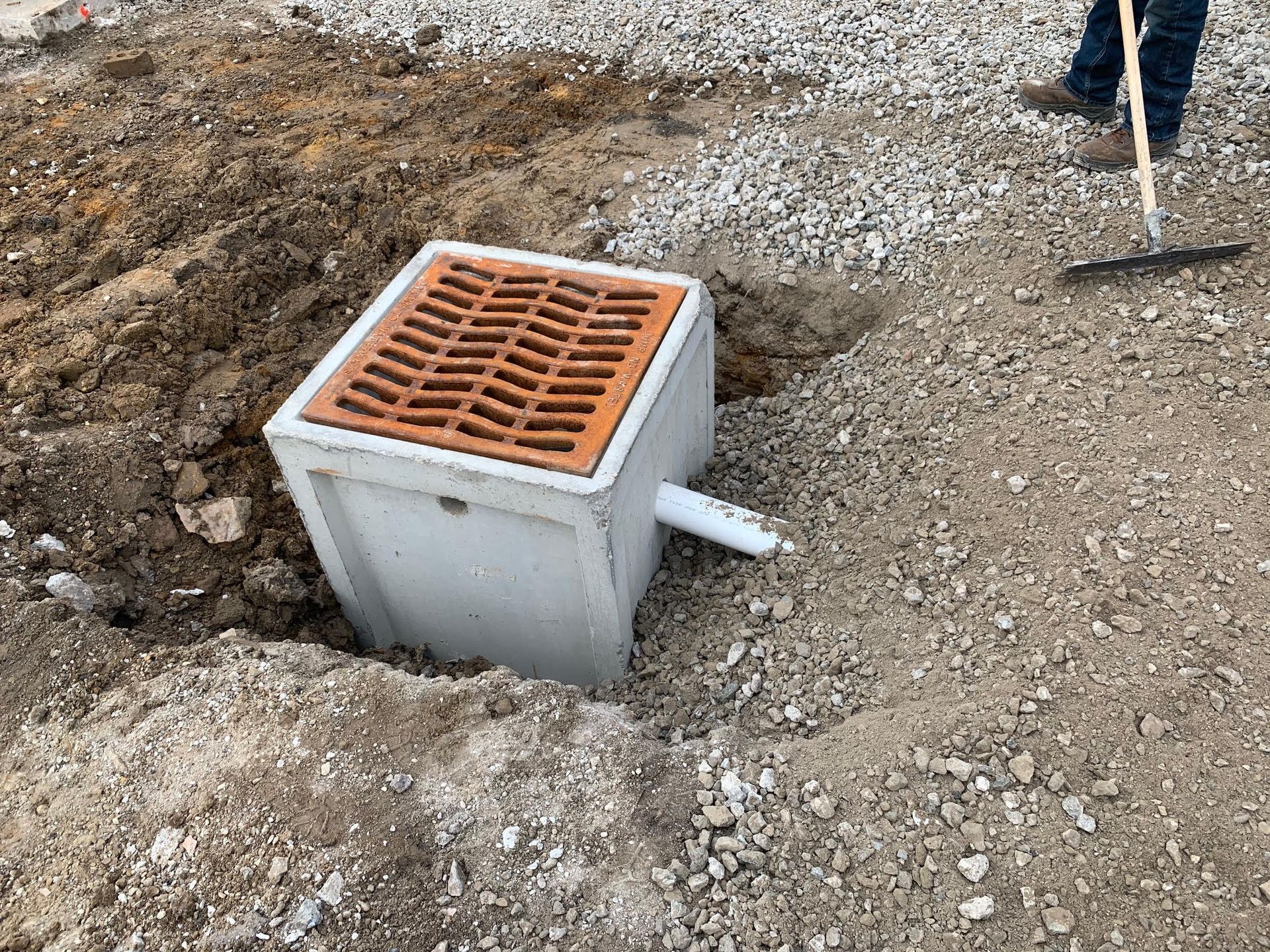Drainage basin with a brick-colored grate surrounded by dirt and gravel. A person is raking in the background.