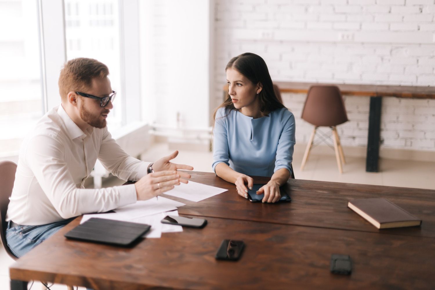 Man gestures while speaking to woman at a table. Papers, phones, and a notebook are on the table.