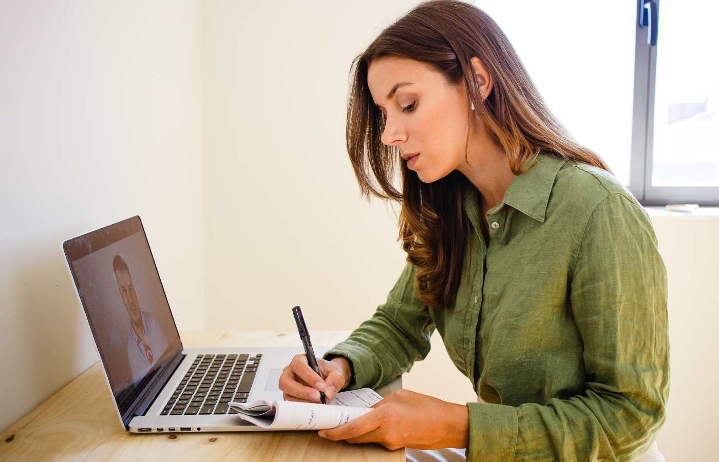 A woman is writing on a piece of paper in front of a laptop computer.