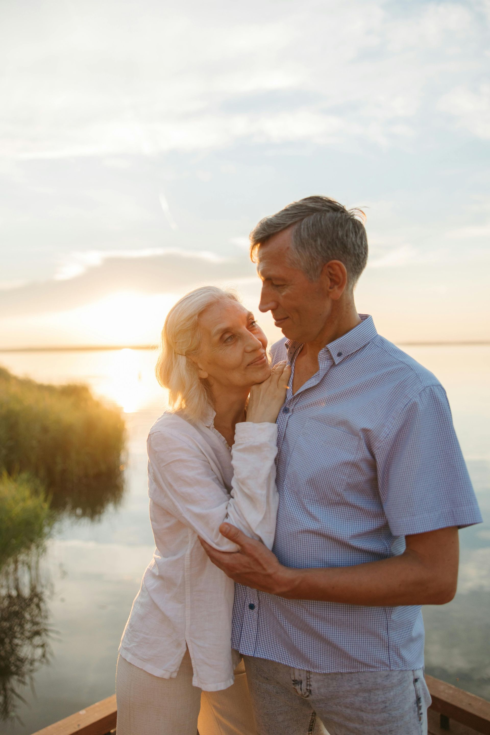 An elderly couple is standing next to each other on a pier near a lake at sunset.