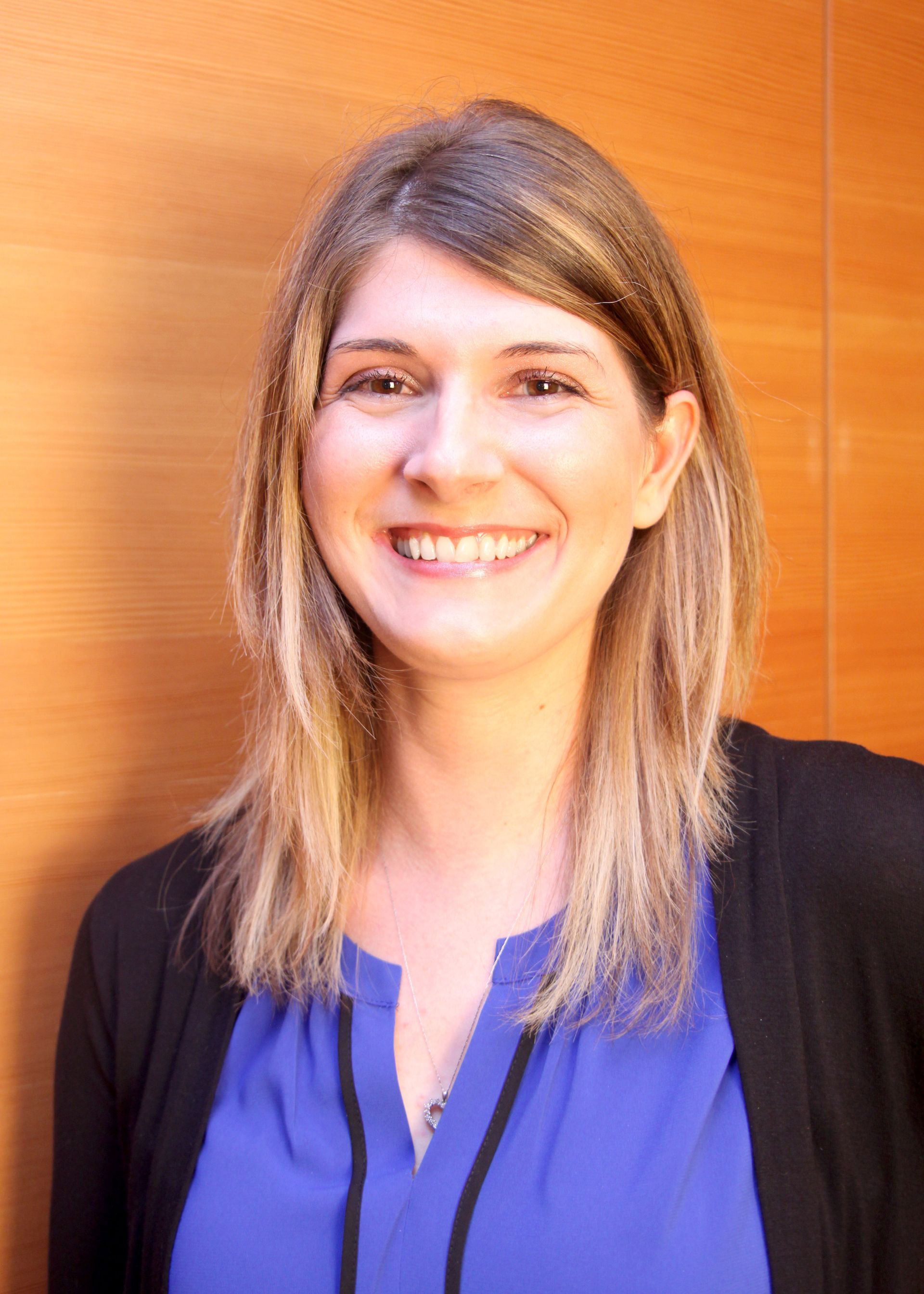 Woman smiling at the camera, wearing a blue shirt and black cardigan; set against a wood panel background.