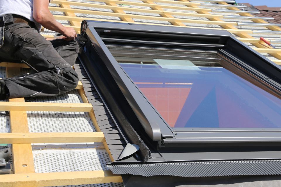 Roofer installing a gray skylight on a wood-framed roof with insulation.