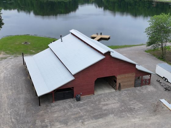 An aerial view of a red barn with a metal roof, located on a gravel lot next to a lake with a wooden dock.