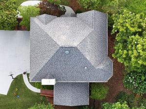 A top-down aerial view of a gray shingled residential roof surrounded by trees and a concrete driveway.