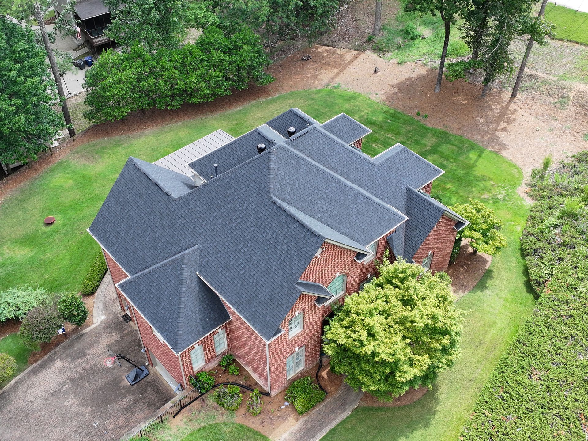 Aerial view of a multi-level red brick house with a dark gray shingled roof, surrounded by a lawn and trees.