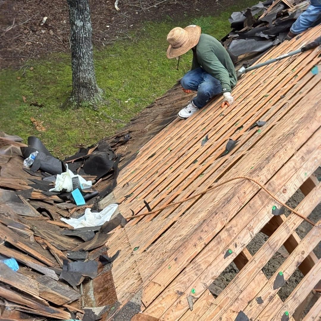 A person wearing a straw hat and green shirt kneeling on a wooden roof frame while removing old shingles.