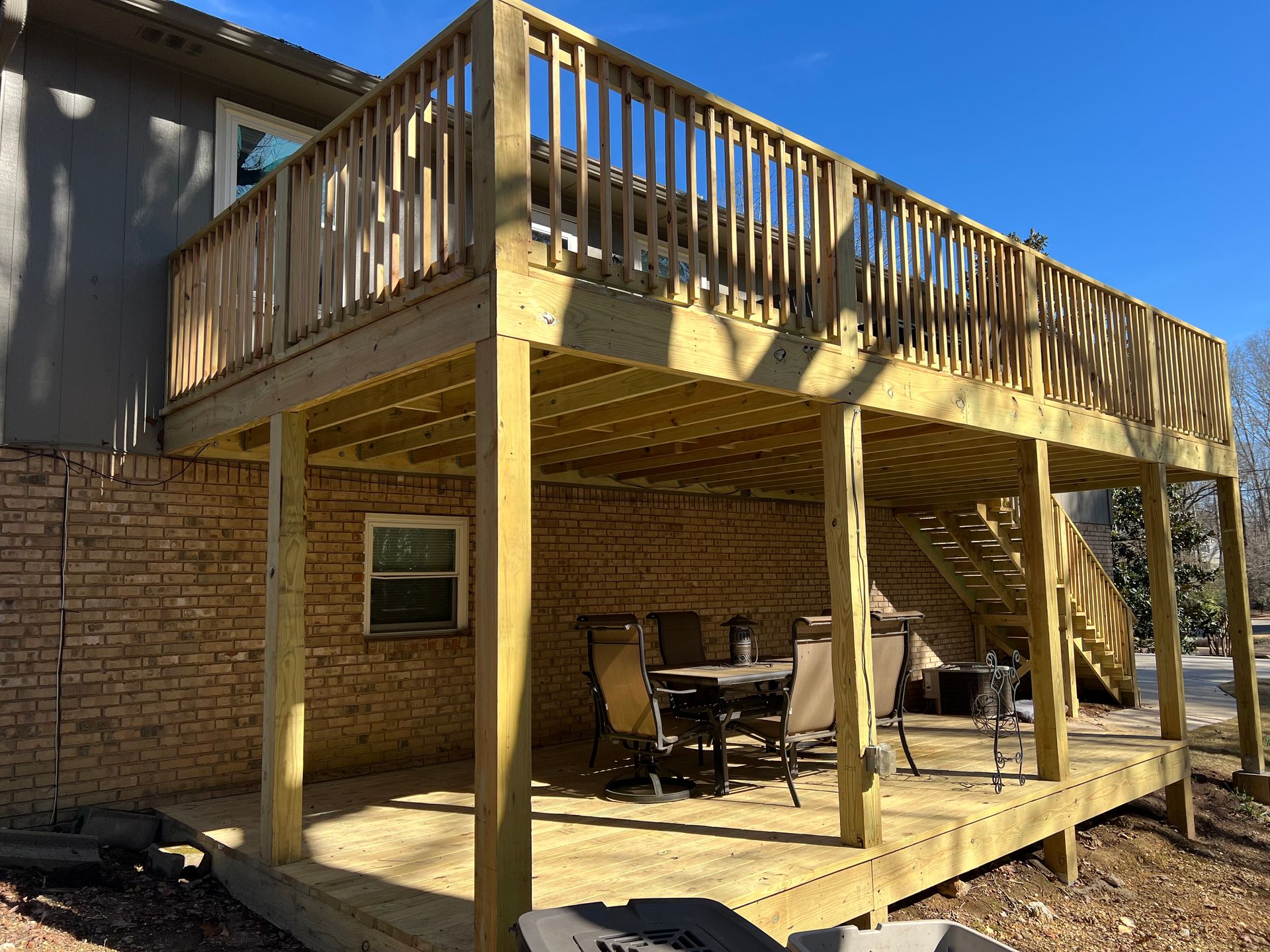 A two-story wooden deck with a stairway, built against the back of a brick house, featuring a lower-level patio area.