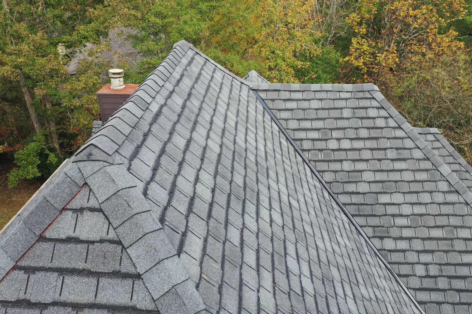 A high-angle view of a gray asphalt shingle roof with a chimney, surrounded by autumn trees.