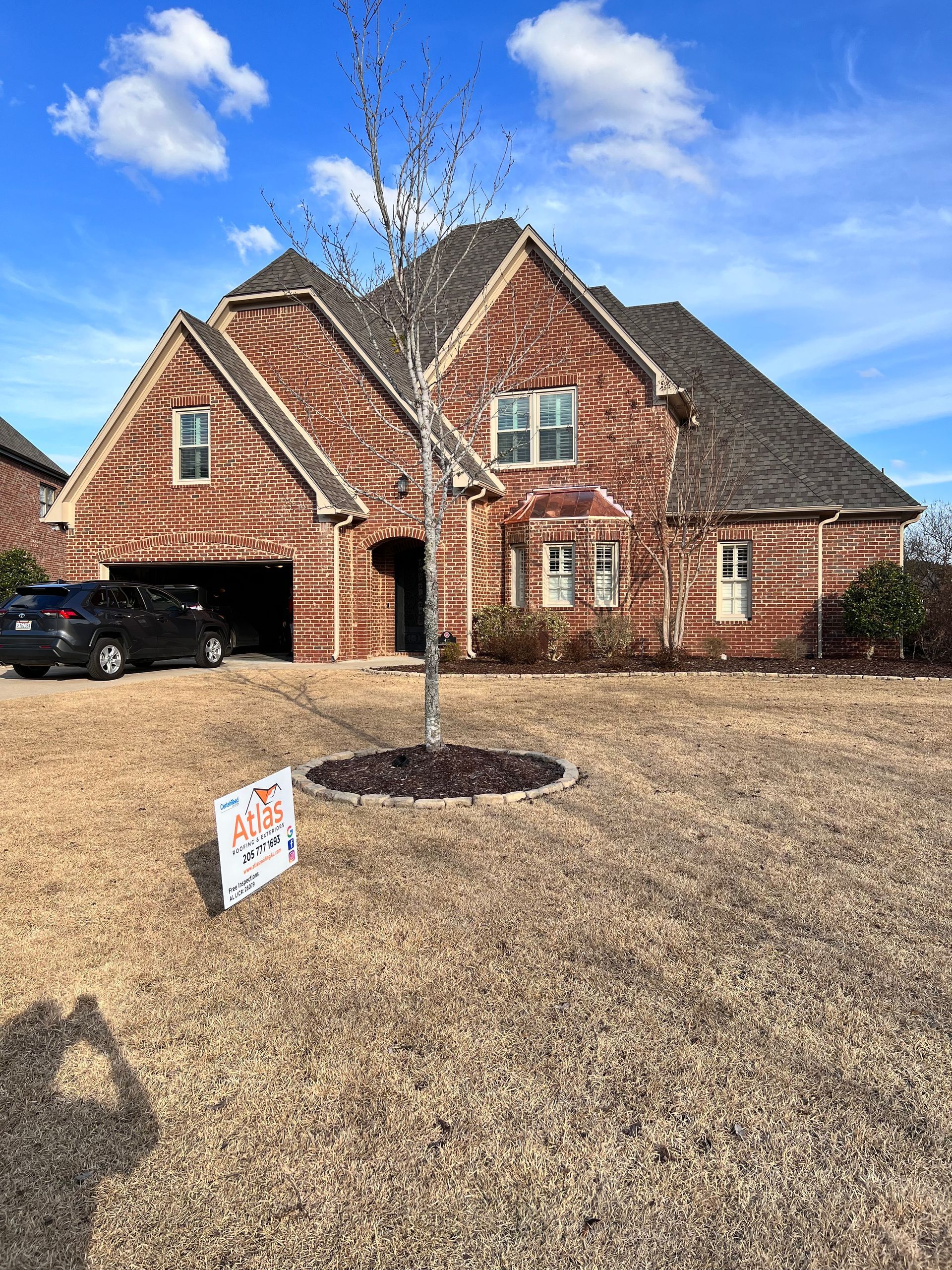 A two-story brick house with a dark roof and a small tree in the front yard, under a blue sky.