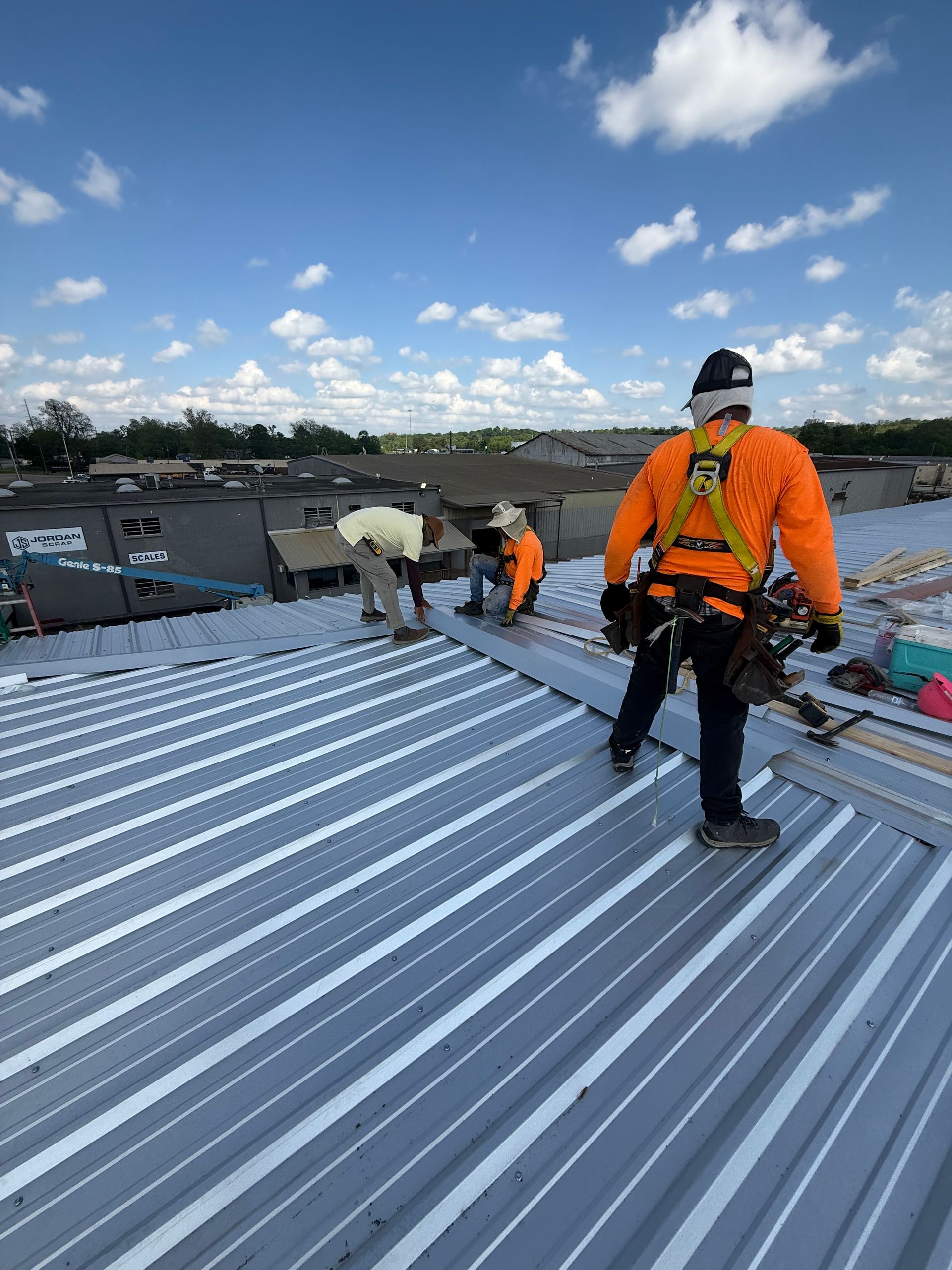 Three workers in safety gear install metal roofing panels on a sunny day.