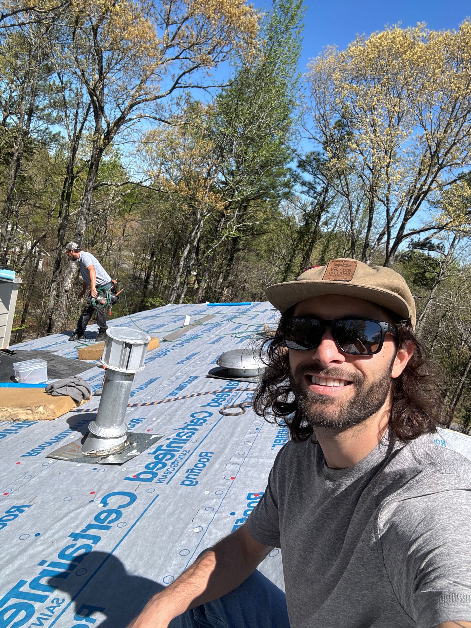 A smiling person in a hat and sunglasses takes a selfie on a roof under construction, with another worker in the back.