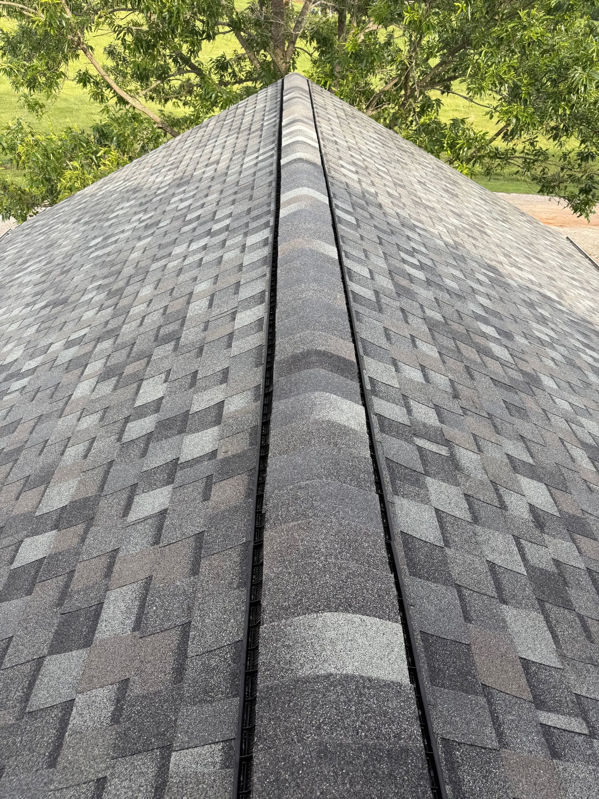 A high-angle view looking down the peak of a roof with gray asphalt shingles and a central ridge cap vent.