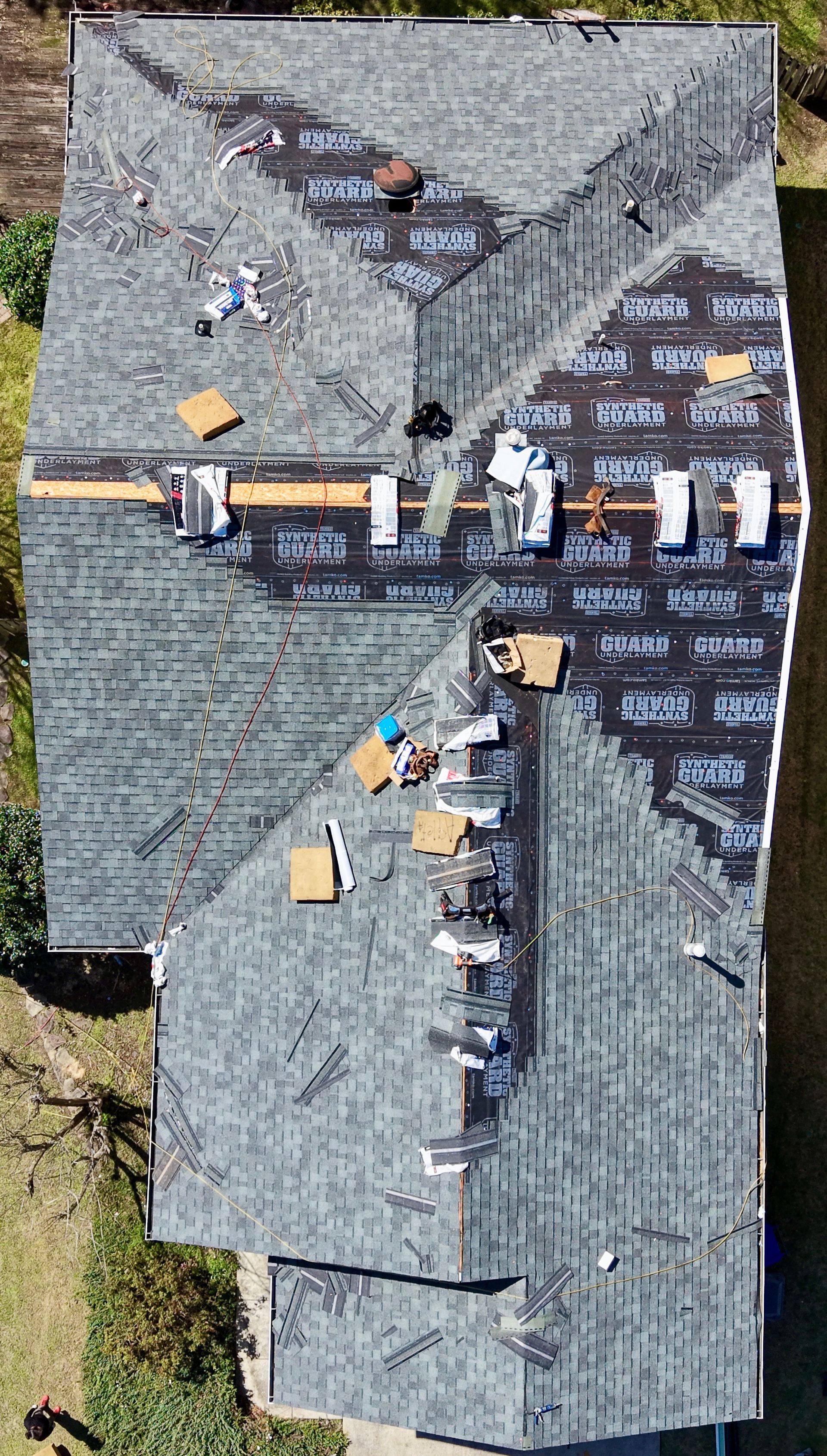 An aerial view of a residential house roof under repair, showing shingles, exposed black underlayment, and roof vents.