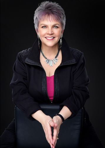 Smiling person seated against a black background, wearing a dark jacket and pink top with a silver necklace.