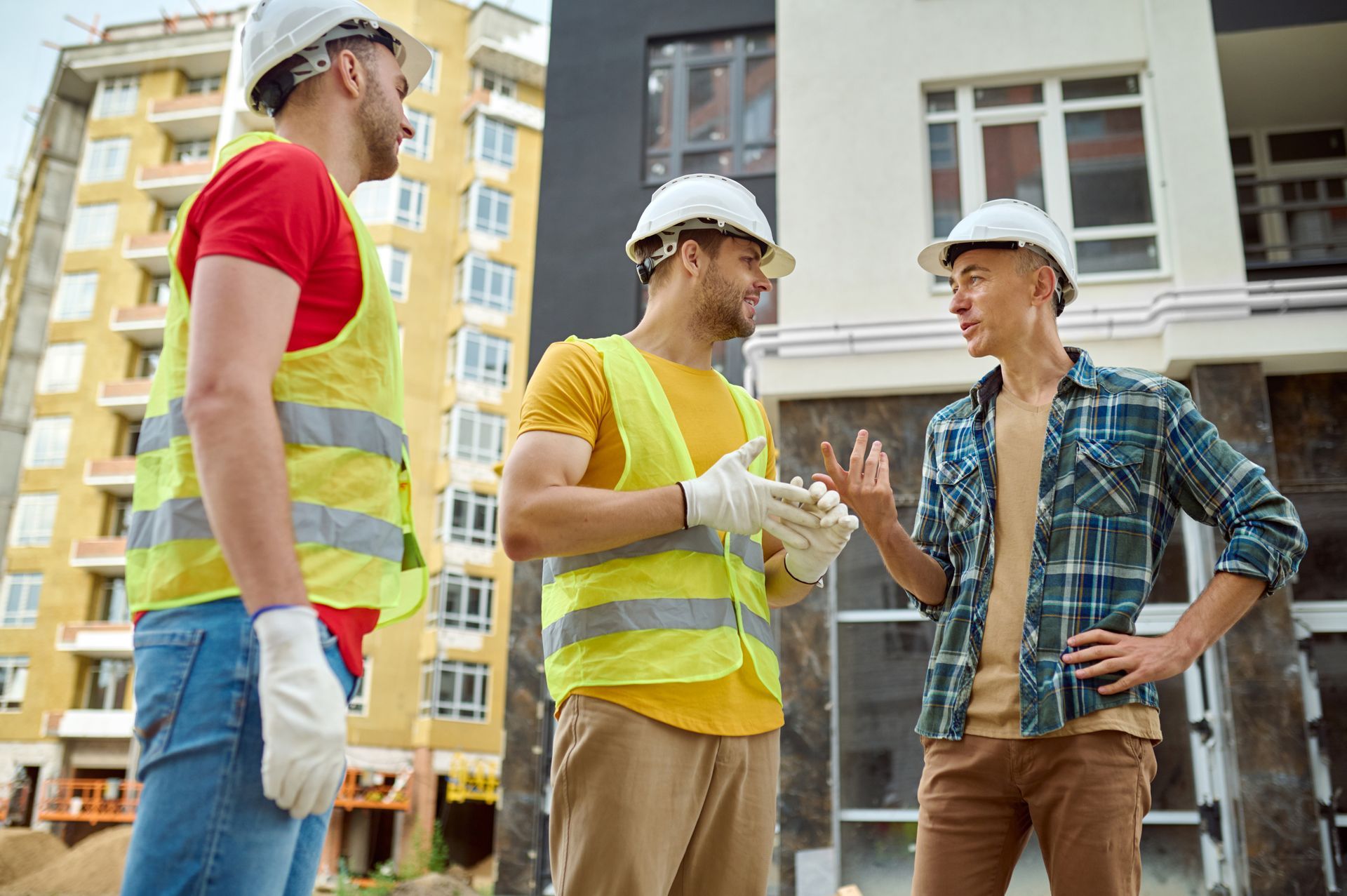 Three construction workers in hard hats and safety vests talking at a building site
