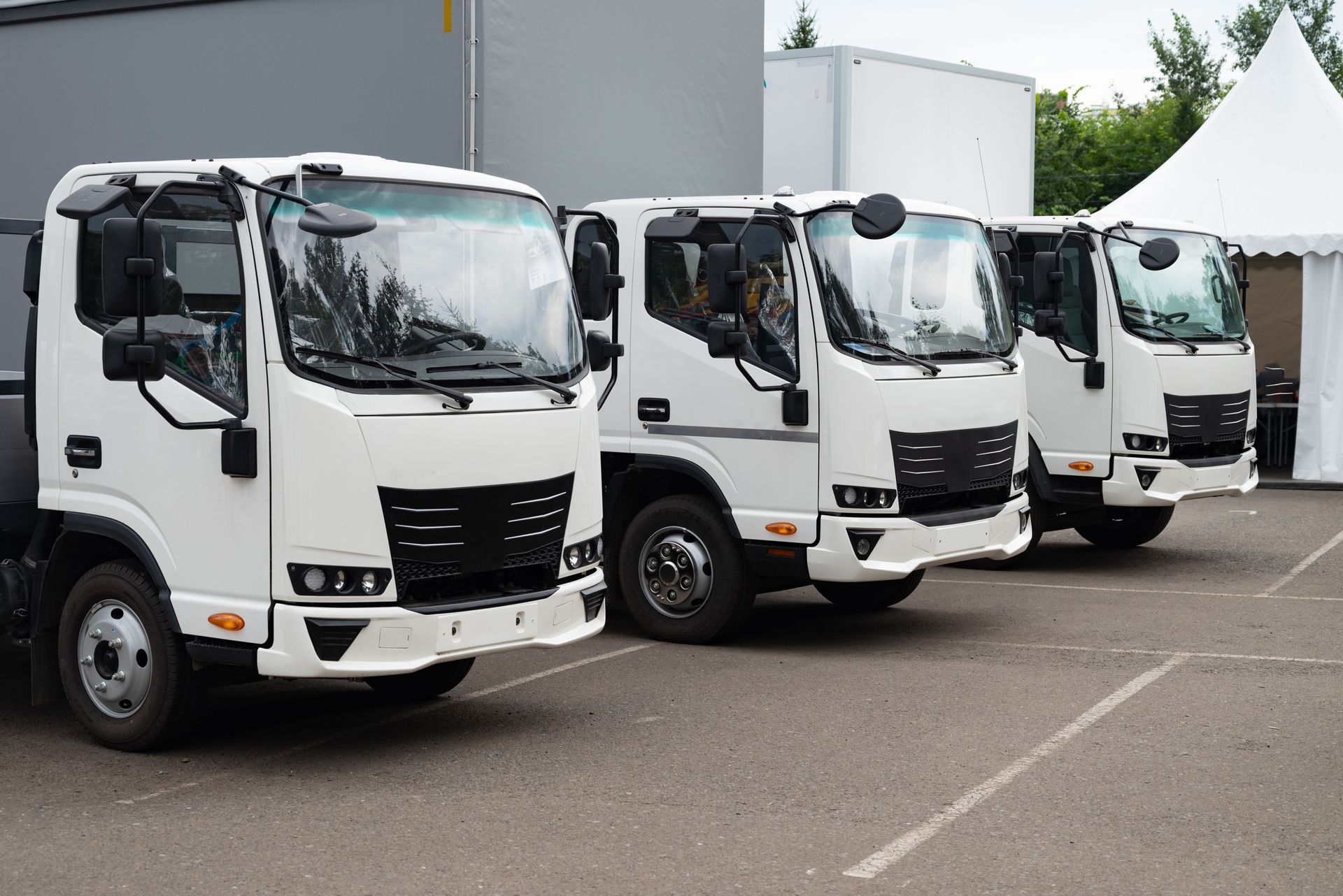 Three white cargo trucks parked in a row on a paved lot near a gray building