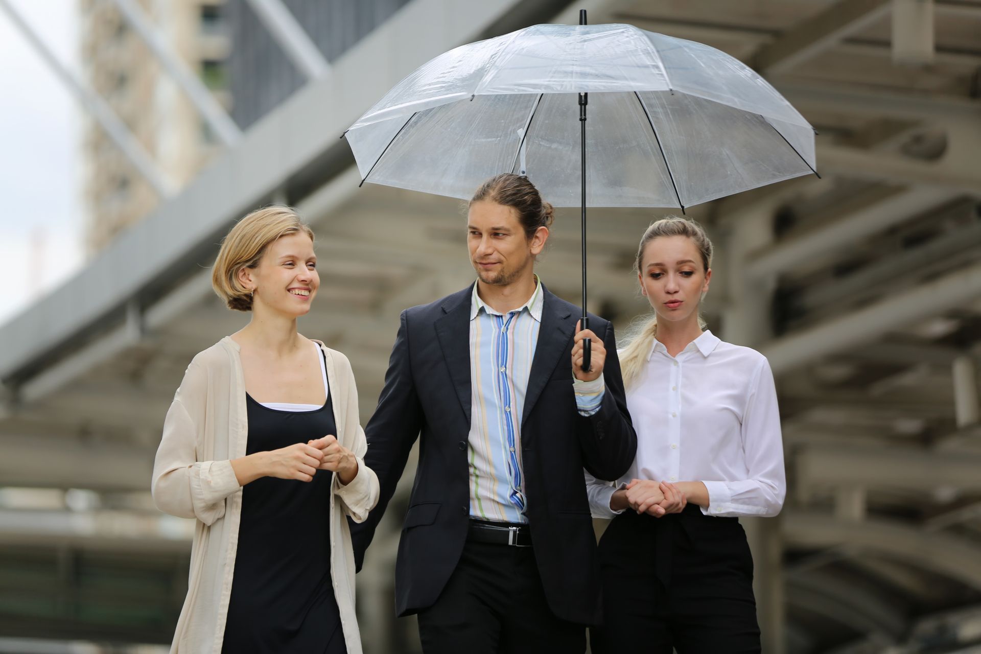 Three coworkers walking under a clear umbrella in an outdoor urban setting