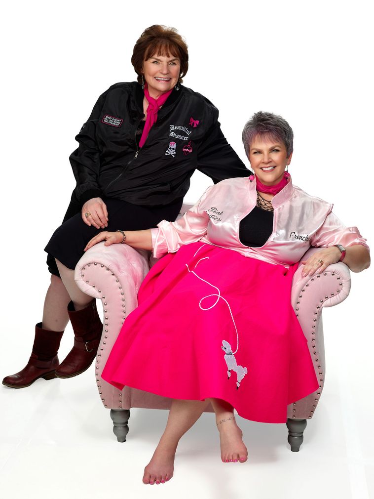 Two women posing in pink chairs, one in black jacket, one in bright pink dress, on a white background