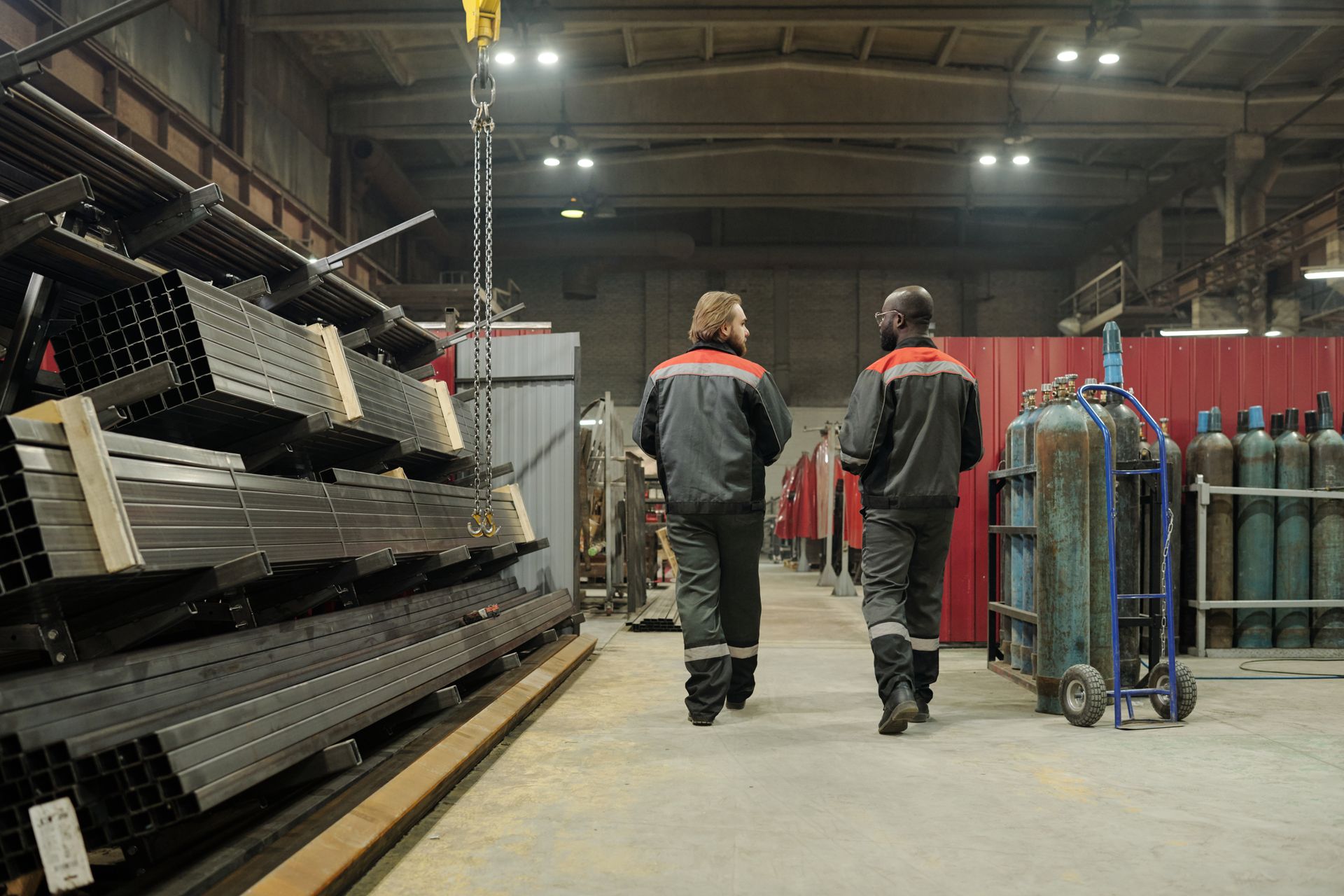 Two workers walking through an industrial warehouse beside stacked metal equipment and carts.
