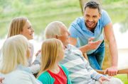 Group of people smiling and talking outdoors on a sunny day, with one man leaning forward and gesturing.