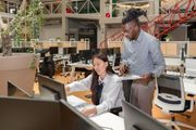 Two coworkers reviewing a computer screen in an open-plan office with desks and monitors.