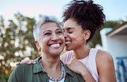 Two smiling women outdoors, one hugging the other from behind at sunset.