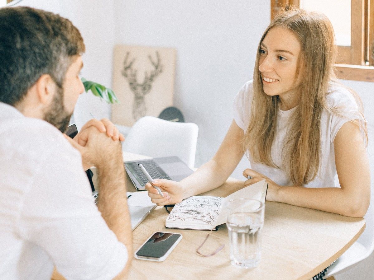 Two people talking at a table in a bright office, with a laptop, phone, notebook, and glass of water.