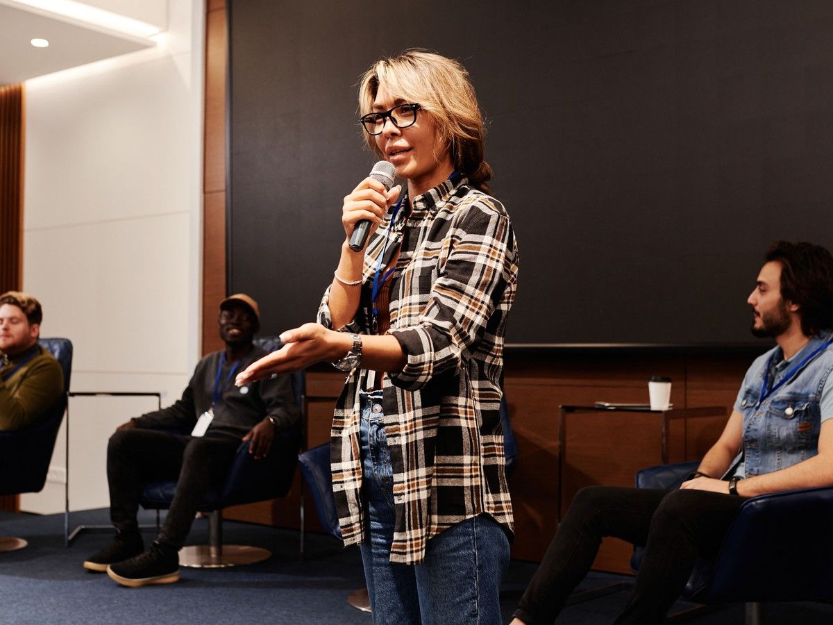 Woman speaking into a microphone at an indoor event, with seated audience members behind her.
