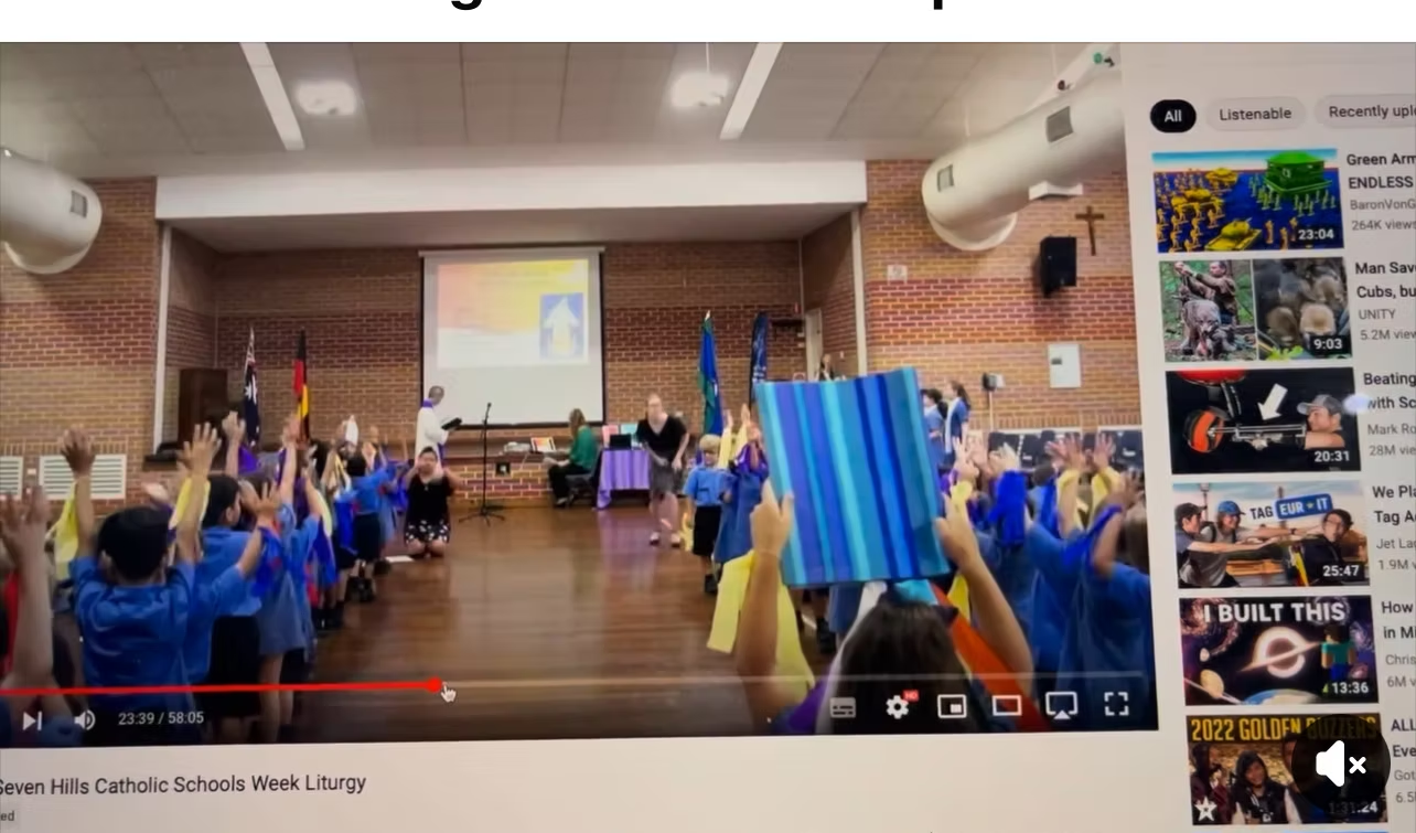 Children in blue uniforms raise hands in a school auditorium. A presentation is on a screen.