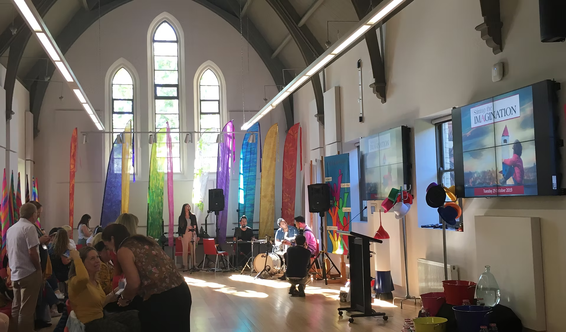 Event in a high-ceilinged room with arched windows; audience, stage with band, screens, colorful flags, and decorations.