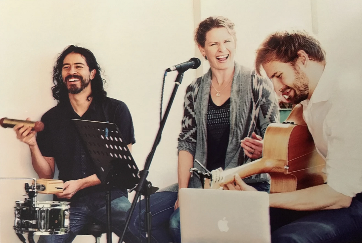 Three musicians laughing, performing. Man plays guitar, man plays percussion, woman sings into a microphone.