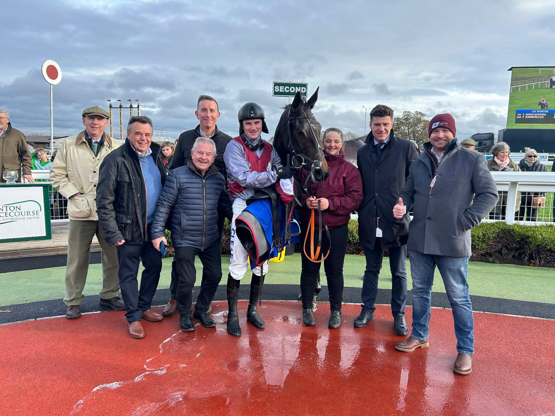 Group posing with a horse and rider on a wet racetrack, with trophy and cloudy sky.
