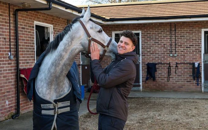 Nick Scholfield offering food to a bay horse in a stable doorway. Brick wall and white window frame.