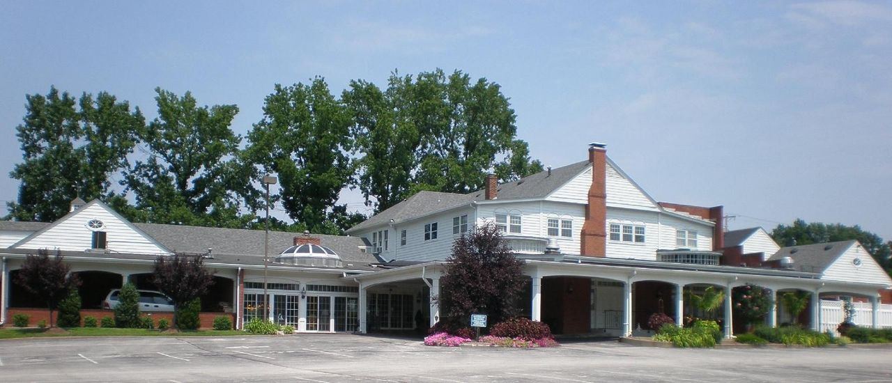 Single-story white storefronts with a red-brick chimney, trees, and a parking lot in front
