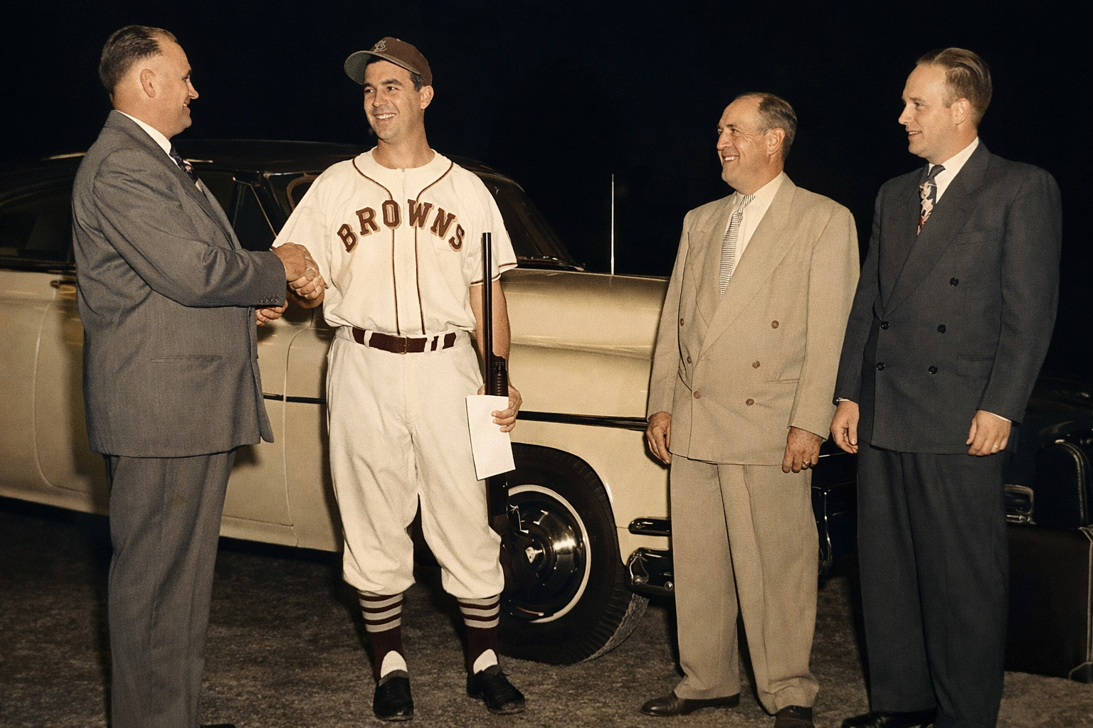 Baseball player in Browns uniform shaking hands with man beside a car while two others look on at night