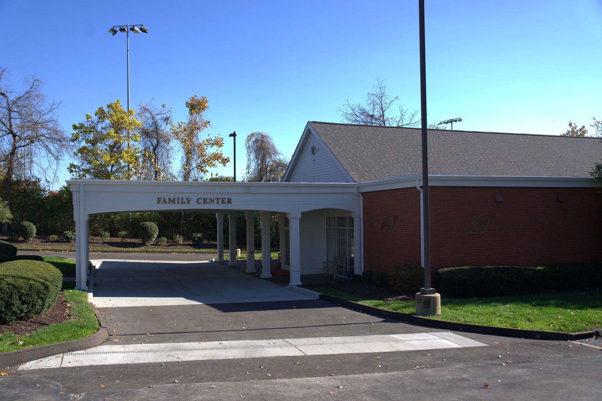 Covered entrance to a brick building with white columns, driveway, and parking lot under a clear blue sky