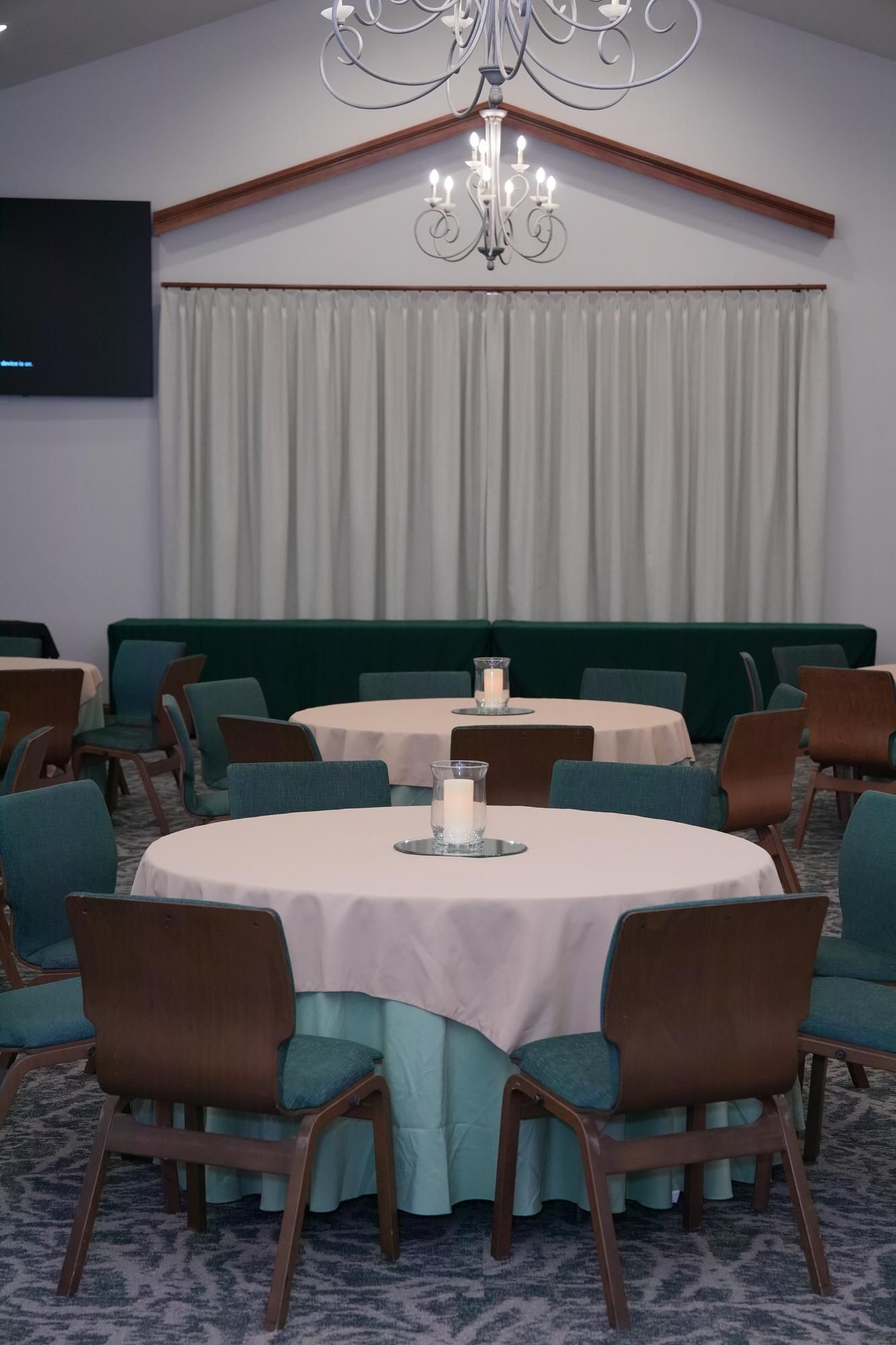 Round banquet tables with white tablecloths in a curtained event hall, with teal chairs and chandeliers.