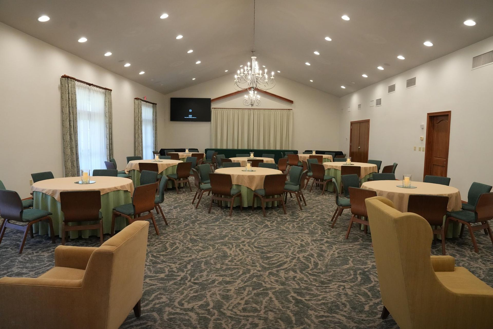 Empty banquet room with round tables, beige chairs, patterned carpet, and chandelier lighting