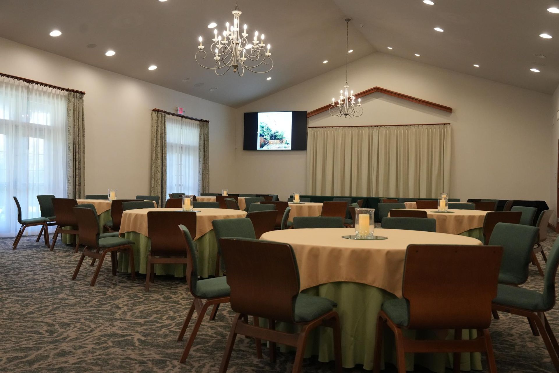 Empty banquet room with round tables, green chair covers, chandeliers, and a wall-mounted TV