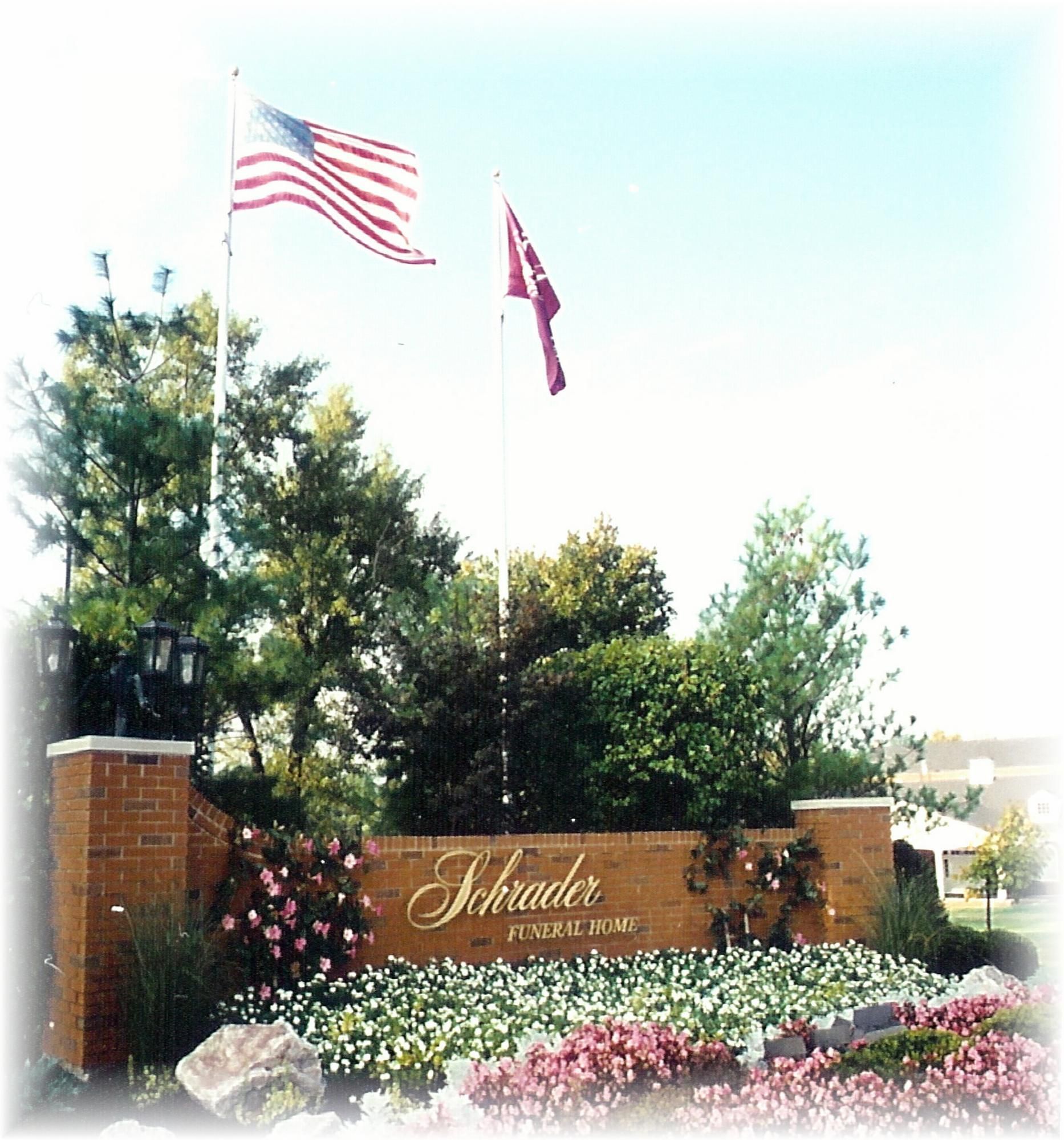 Subdivision entrance sign with flowers, trees, and two flags in front of a brick wall